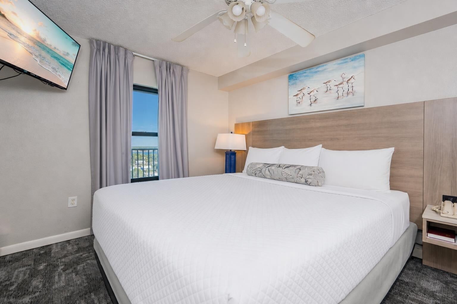 A hotel room with a large bed, a ceiling fan, a painting, a nightstand, and a view of the outdoors.