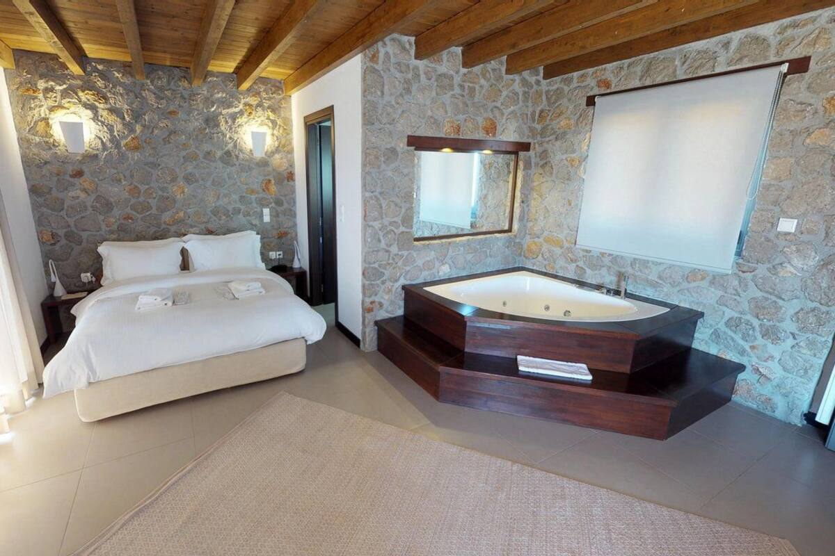 Een slaapkamer met een stenen muur, een groot bed, een houten plafond en een moderne badkamer met een vrijstaand bad.