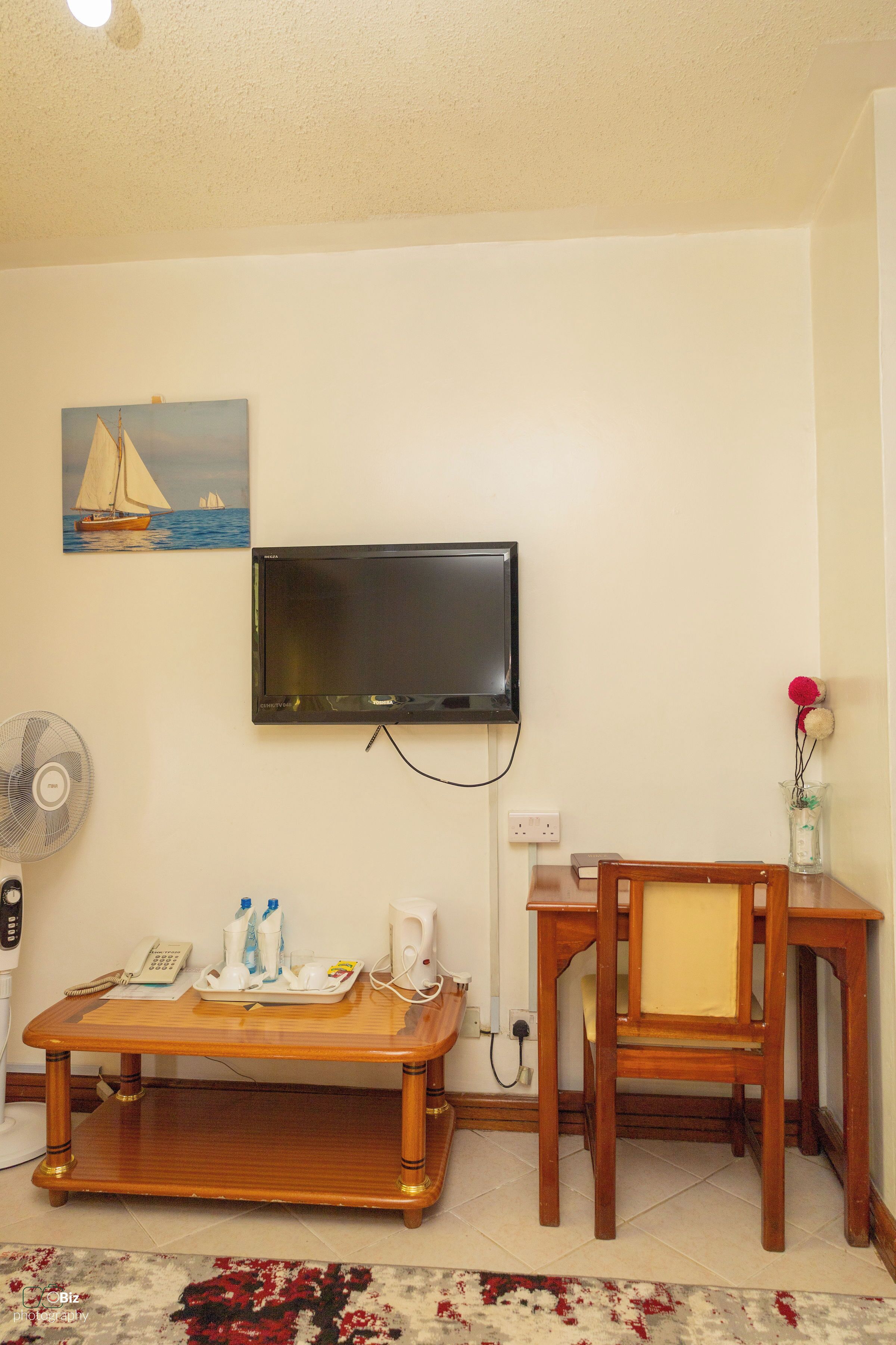 Un salón con una televisión de pantalla plana montada en la pared, una mesa de centro de madera con varios objetos, una mesita de madera con un jarrón y una alfombra estampada en el suelo.