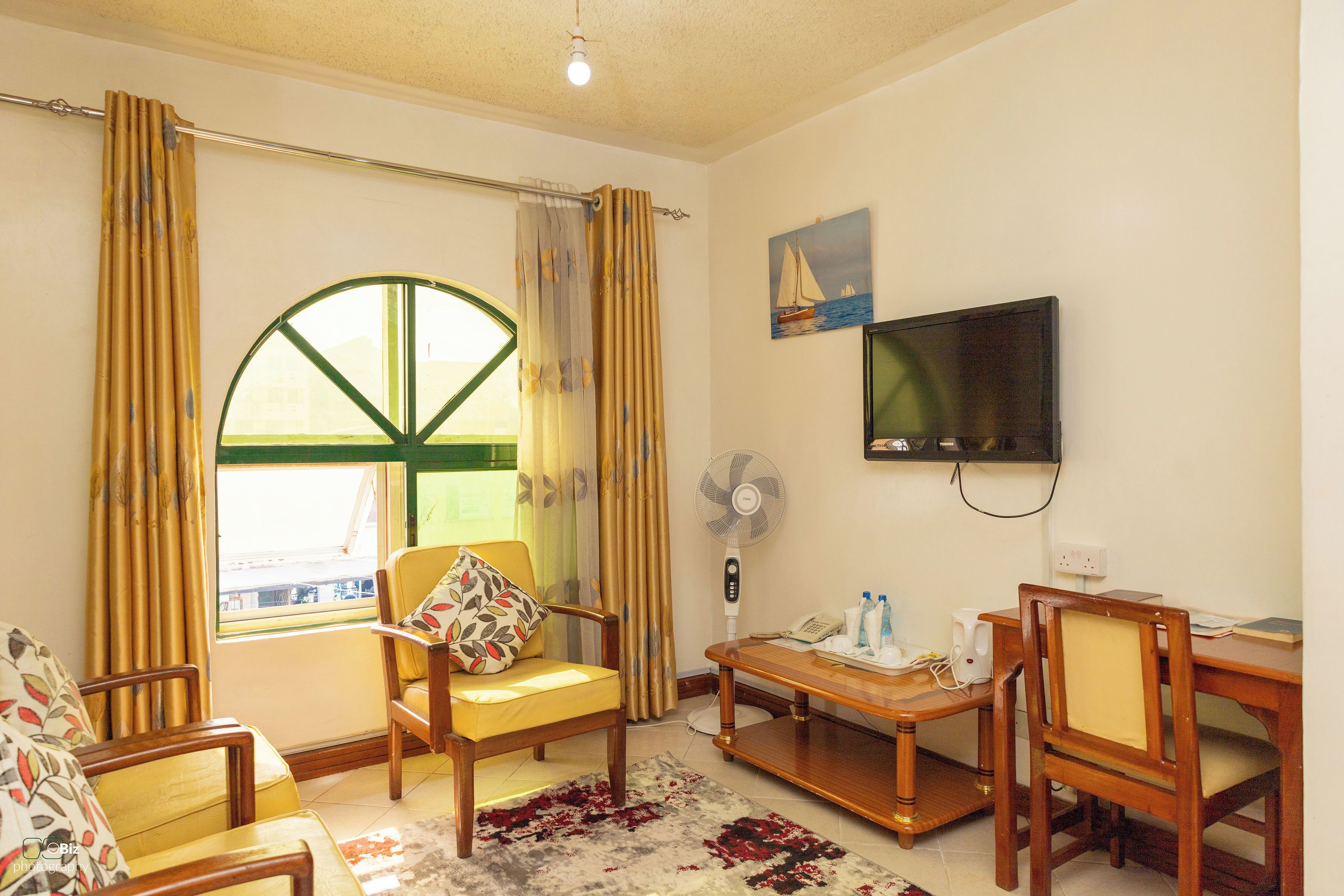 Una sala de estar con un ventanal grande, un televisor montado en la pared, un ventilador y una mesa de madera con sillas.