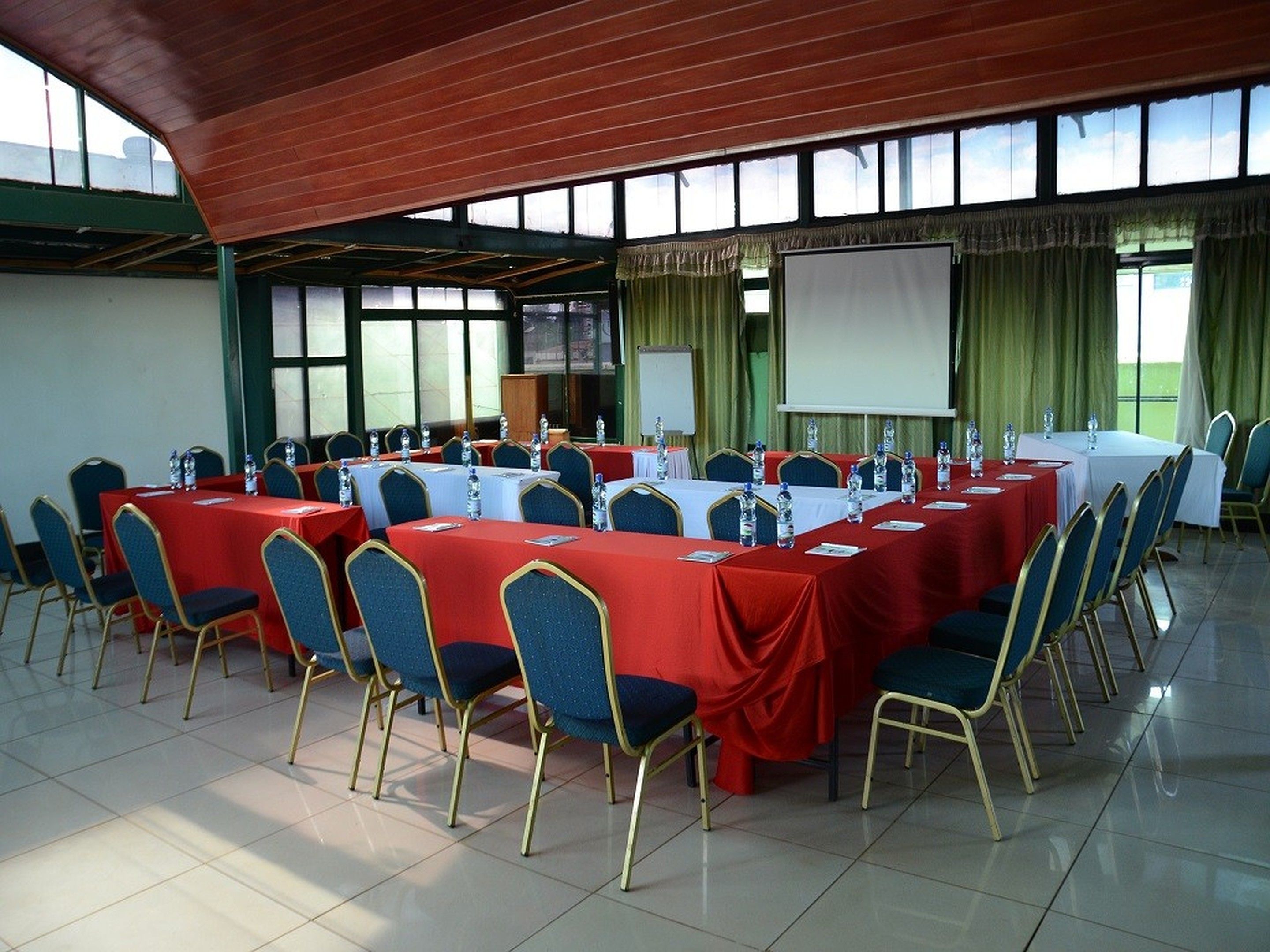 Un salón de conferencias con varias mesas y sillas, ventanas amplias y una pantalla para proyector.