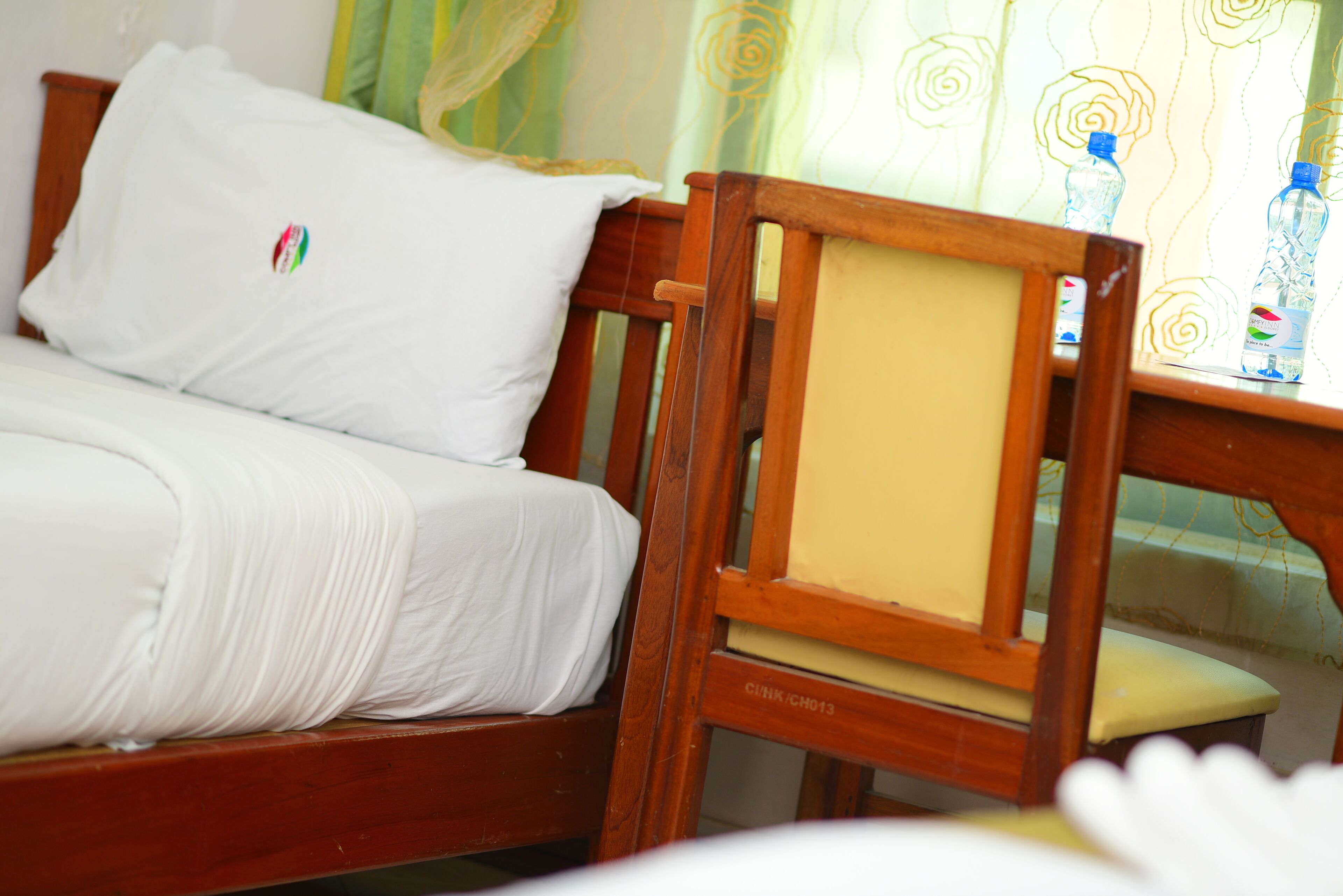 Una cama con ropa de cama blanca y una almohada con un logotipo, una silla de madera con cojín, y una mesa con una botella de agua.