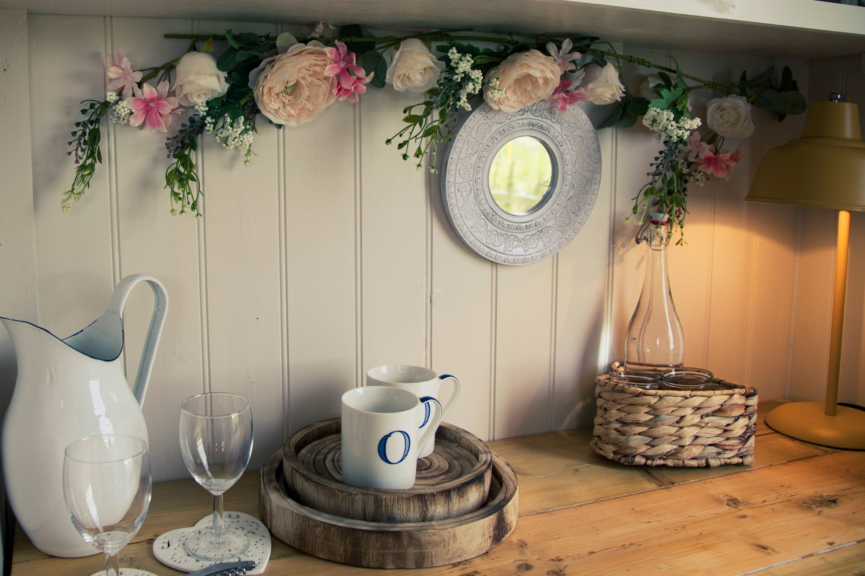A wooden table with a pitcher, two cups, a glass, a woven basket, a mirror, and a vase with flowers.