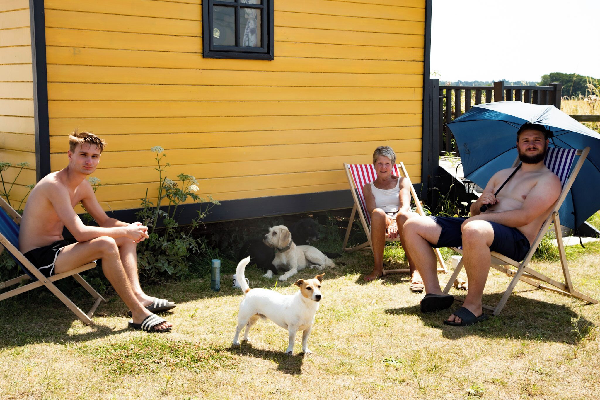 Three people sitting on lawn chairs outside a yellow house with a dog nearby.