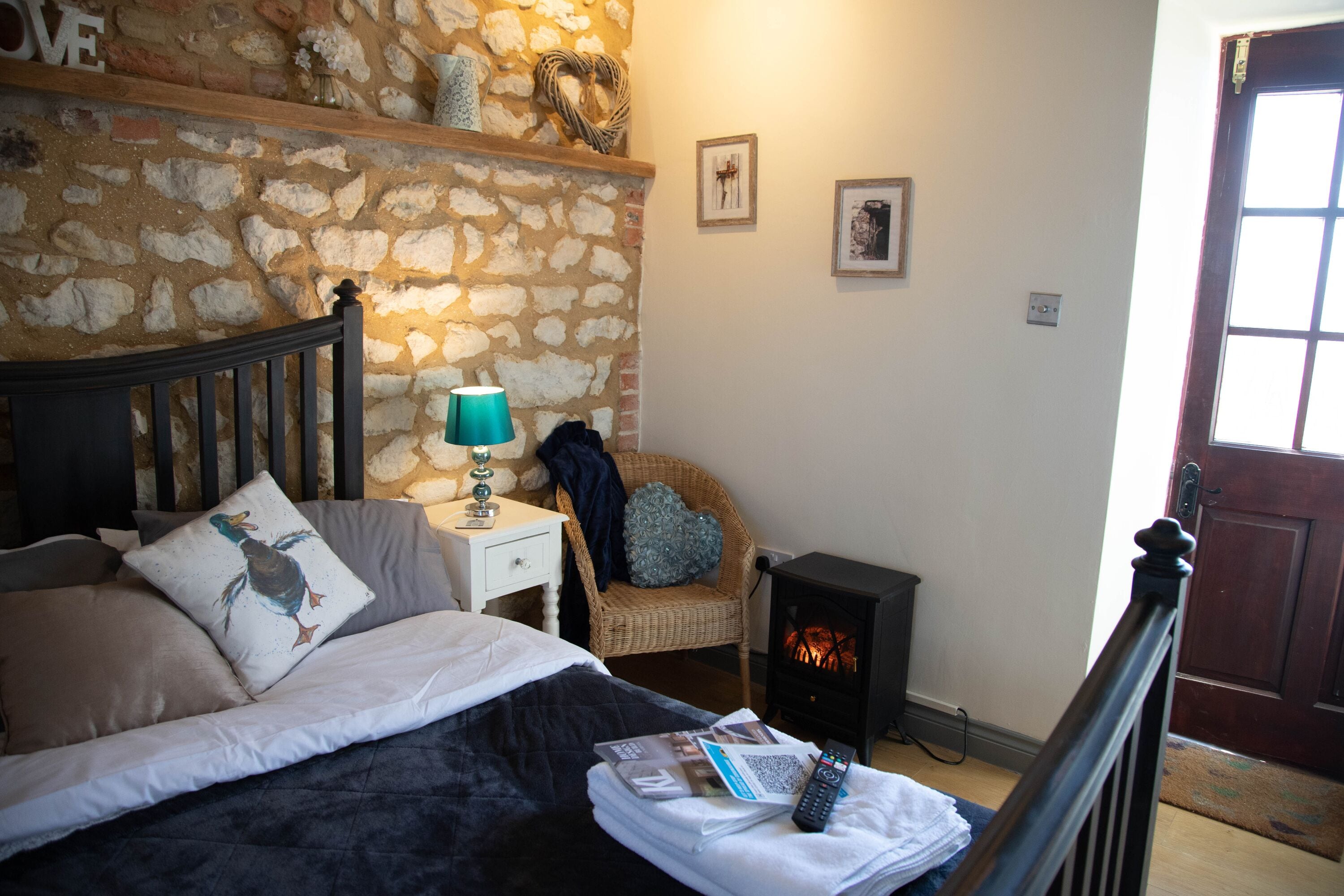 A bedroom with a stone wall, a bed with pillows, a nightstand with a lamp, a chair, and a fireplace.
