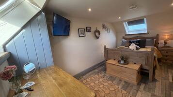 A cozy bedroom with a wooden bed, a small bedside table, a TV mounted on the wall, and a window providing natural light.