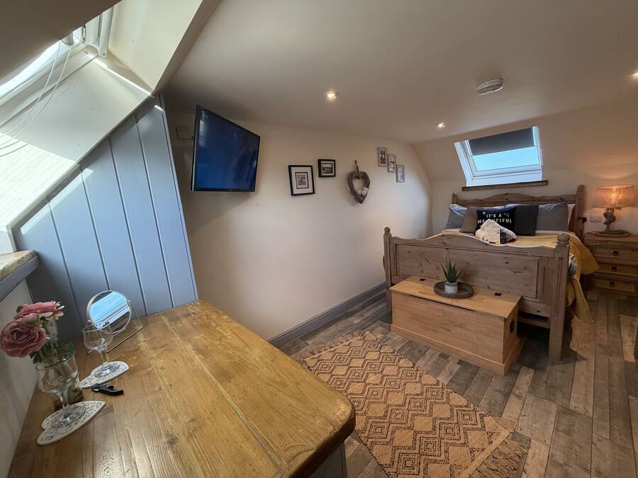 A cozy bedroom with a wooden bed, a small bedside table, a TV mounted on the wall, and a window providing natural light.