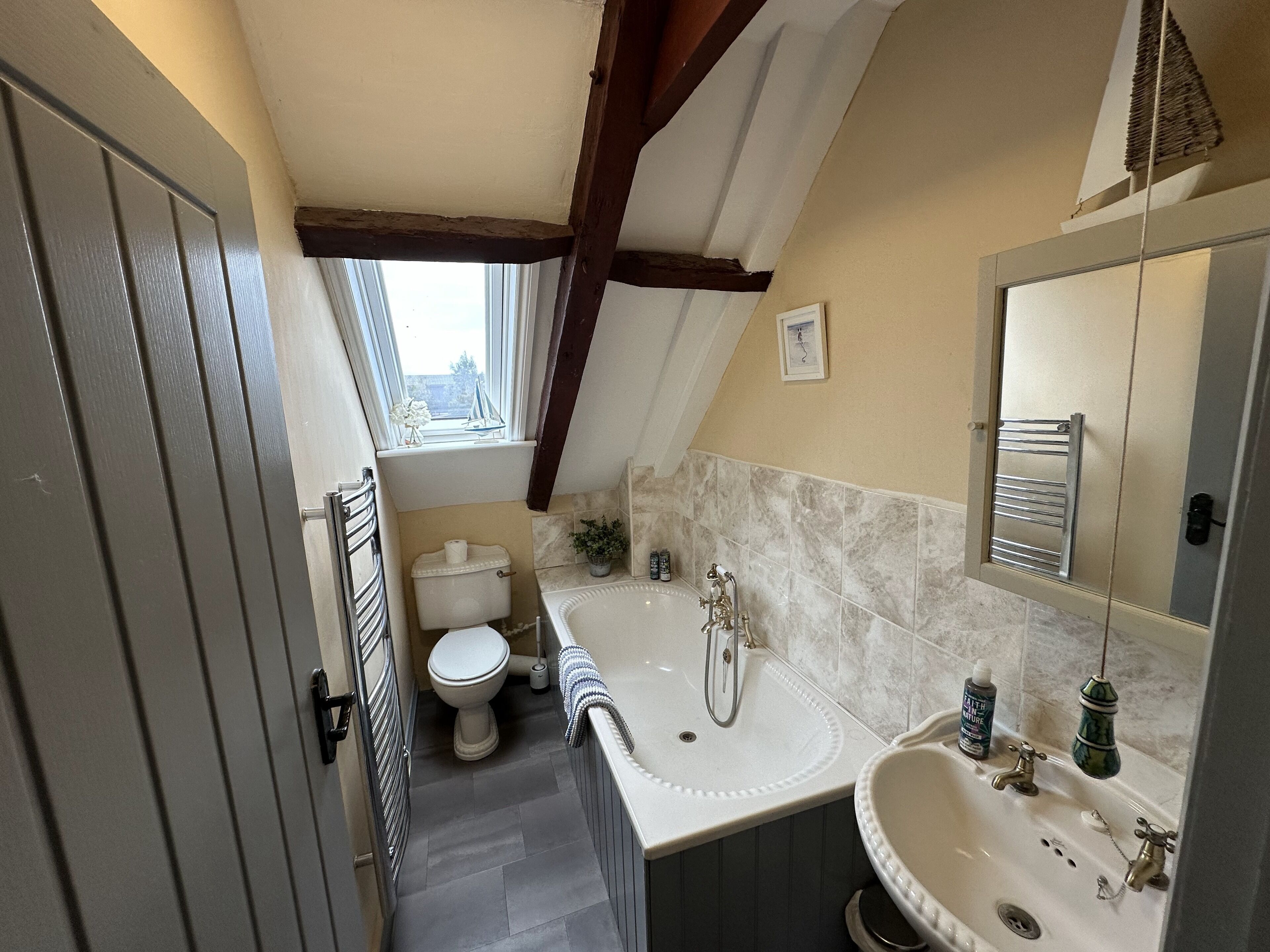 Een kleine, praktische badkamer met toilet, wastafel en bad. Er is een raam voor daglicht, en de muren zijn betegeld.