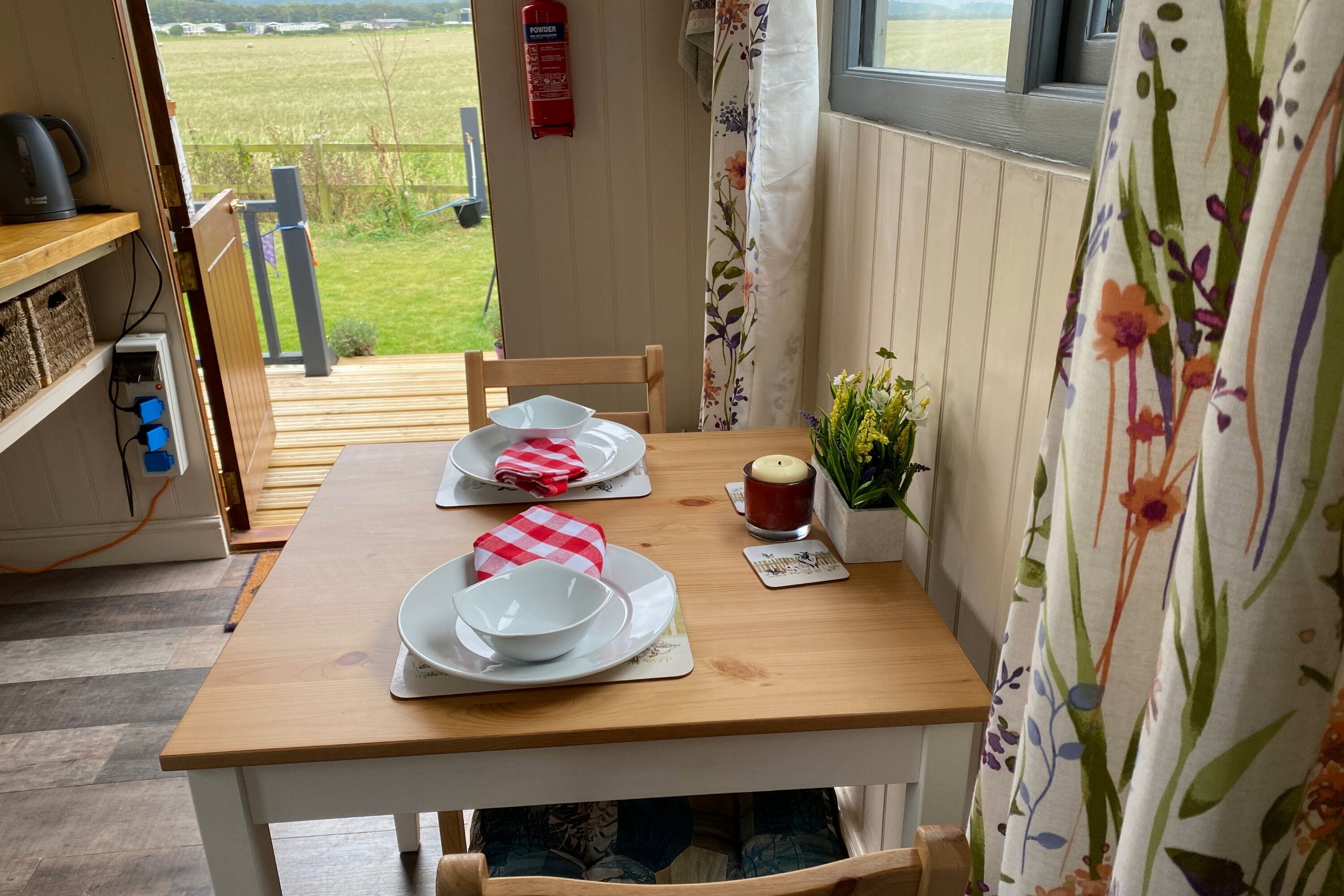 Una mesa de madera con platos, tazones y una vela, frente a una ventana que da a un campo.