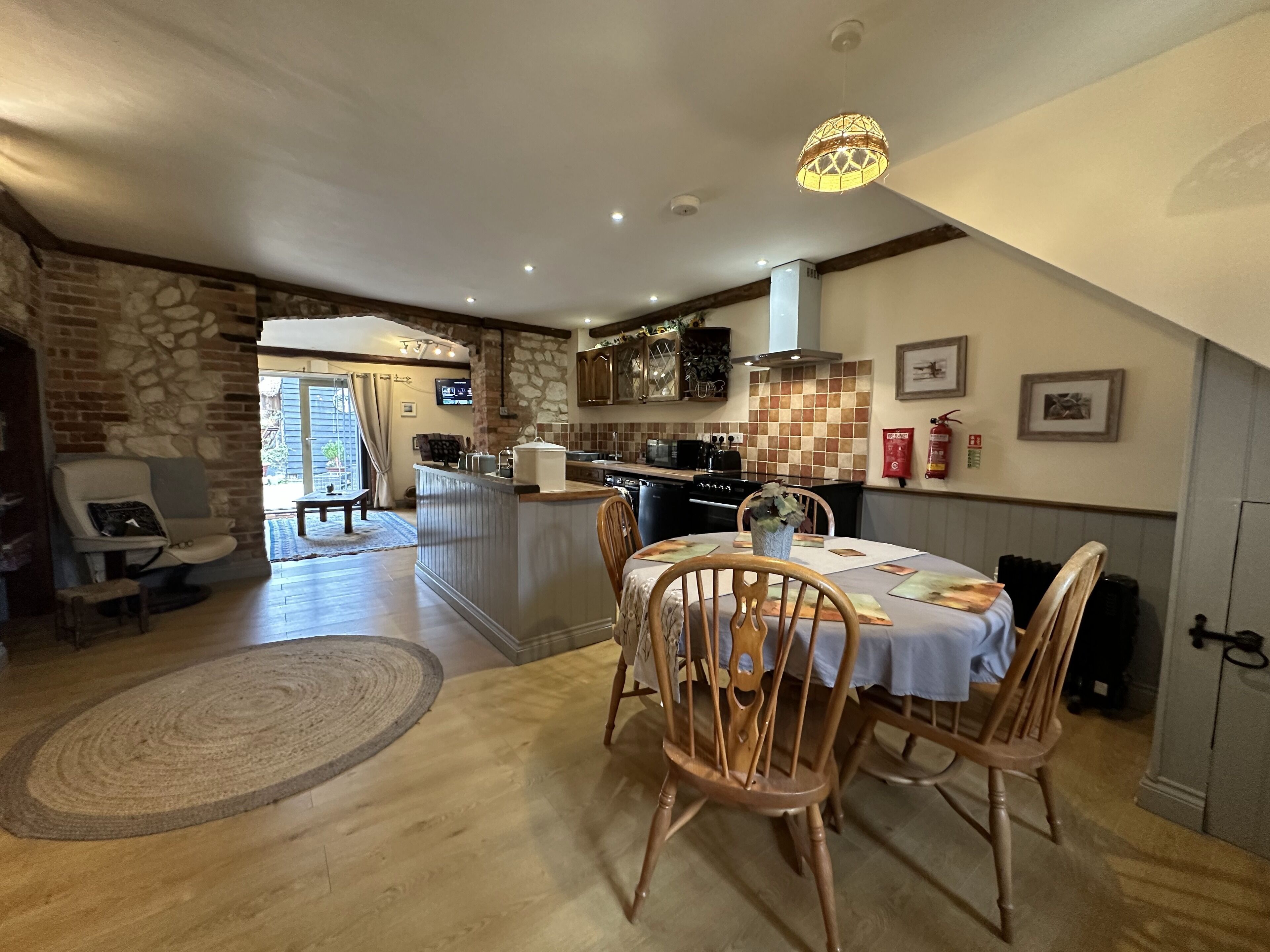 A cozy dining area with a wooden table and chairs, a kitchen in the background, and a stone wall.