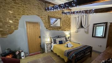 A cozy bedroom with a stone wall, a wooden door, a bed with a yellow blanket, a bedside table with a lamp, and a window with curtains.