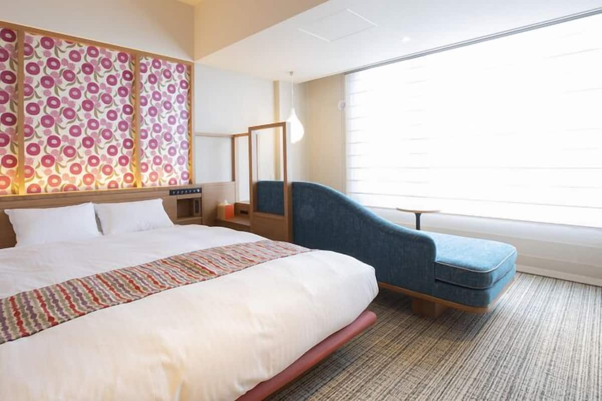 Habitación de hotel con una cama grande, un sillón azul, un papel tapiz estampado y una ventana con persianas.