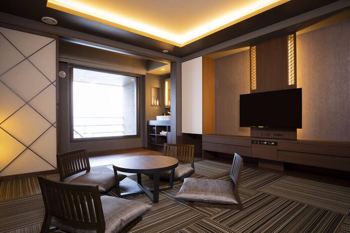 Una habitación de hotel con televisión, una mesa de comedor, sillas, un sofá y un ventanal grande.