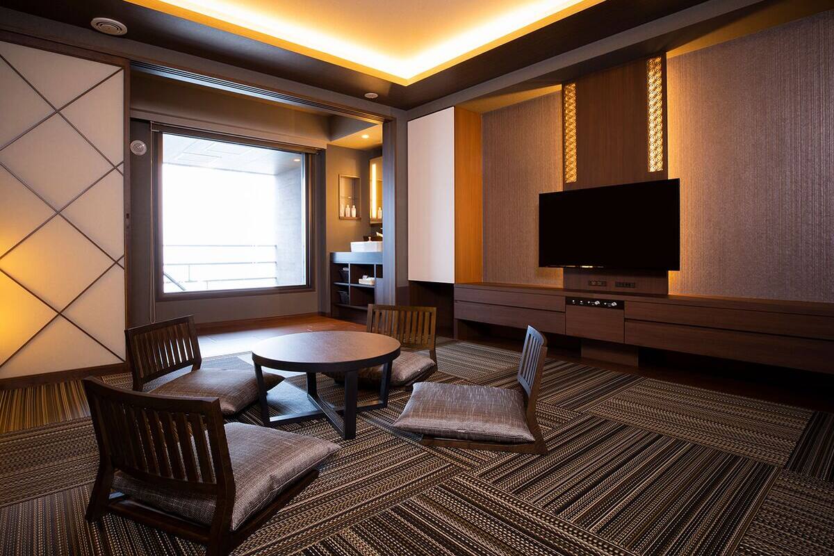 Habitación de hotel con comedor, televisor de pantalla plana y un amplio ventanal que proporciona luz natural.