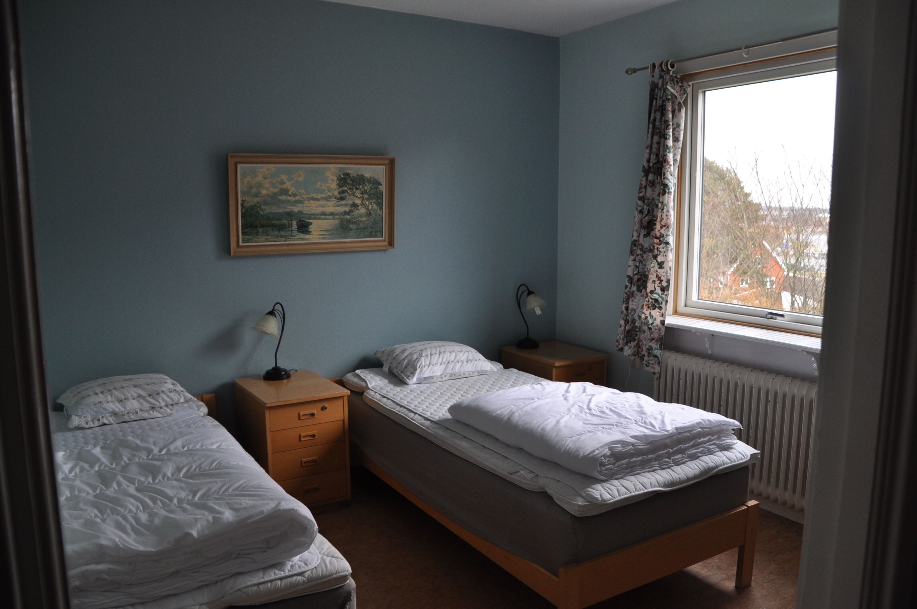A bedroom with two single beds, a nightstand with a lamp, a dresser, and a window with a view of houses.