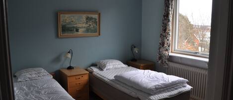 A bedroom with two single beds, a nightstand with a lamp, a dresser, and a window with a view of houses.