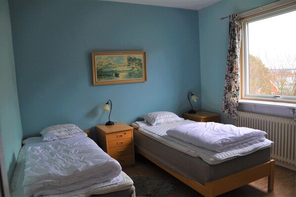 A room with two single beds, a wooden nightstand with a lamp, a framed painting on the wall, and a window with curtains. - Roddaren Vandrarhem - Hostel (Stromstad)