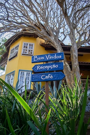 A yellow building with a signpost indicating directions to 'Bem Vindos,' 'Reception,' and 'Cafe.' - Villa Bella Ferradura (Búzios)