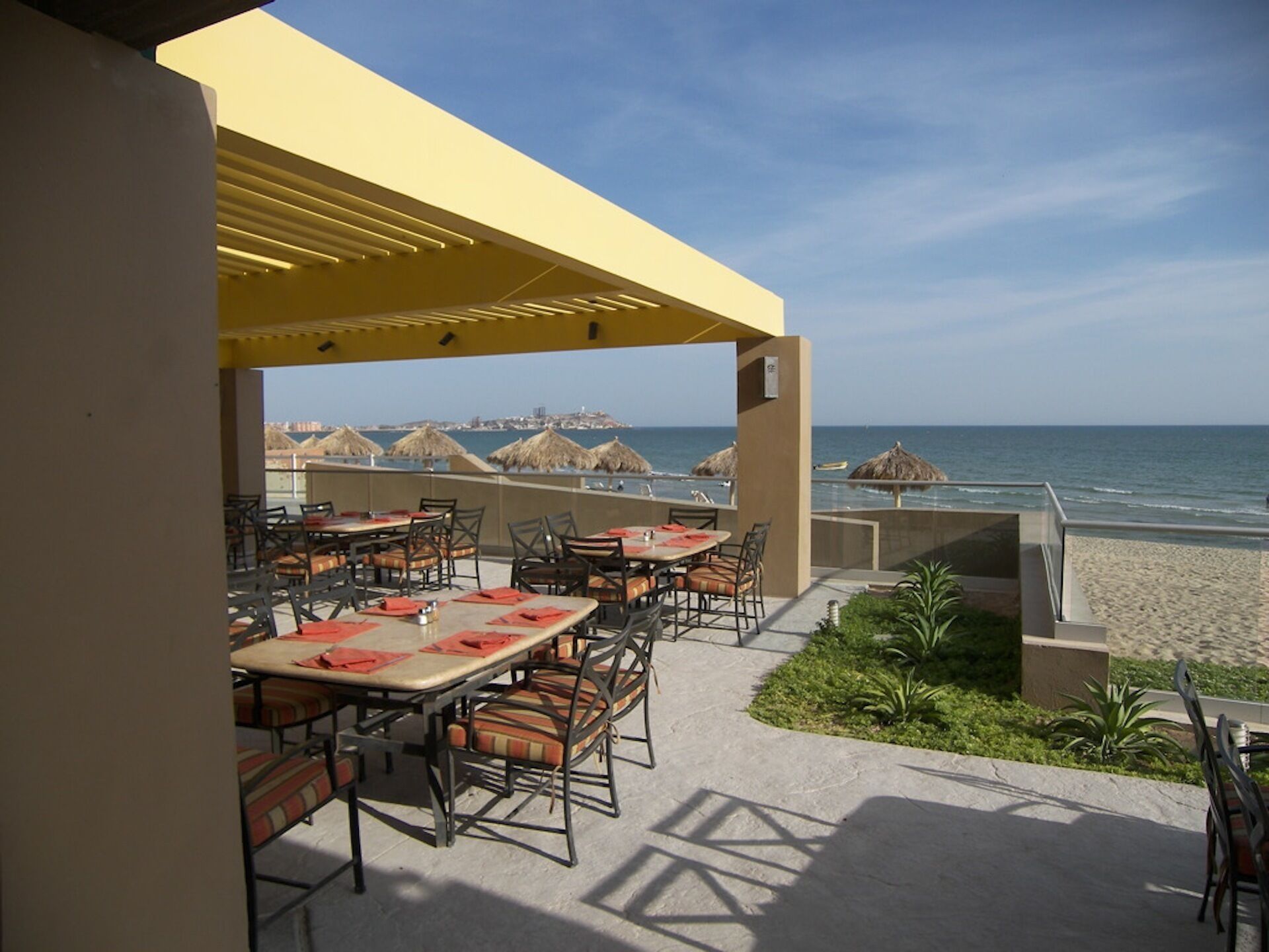 An outdoor dining area with tables and chairs, overlooking a beach with thatched umbrellas and the ocean.