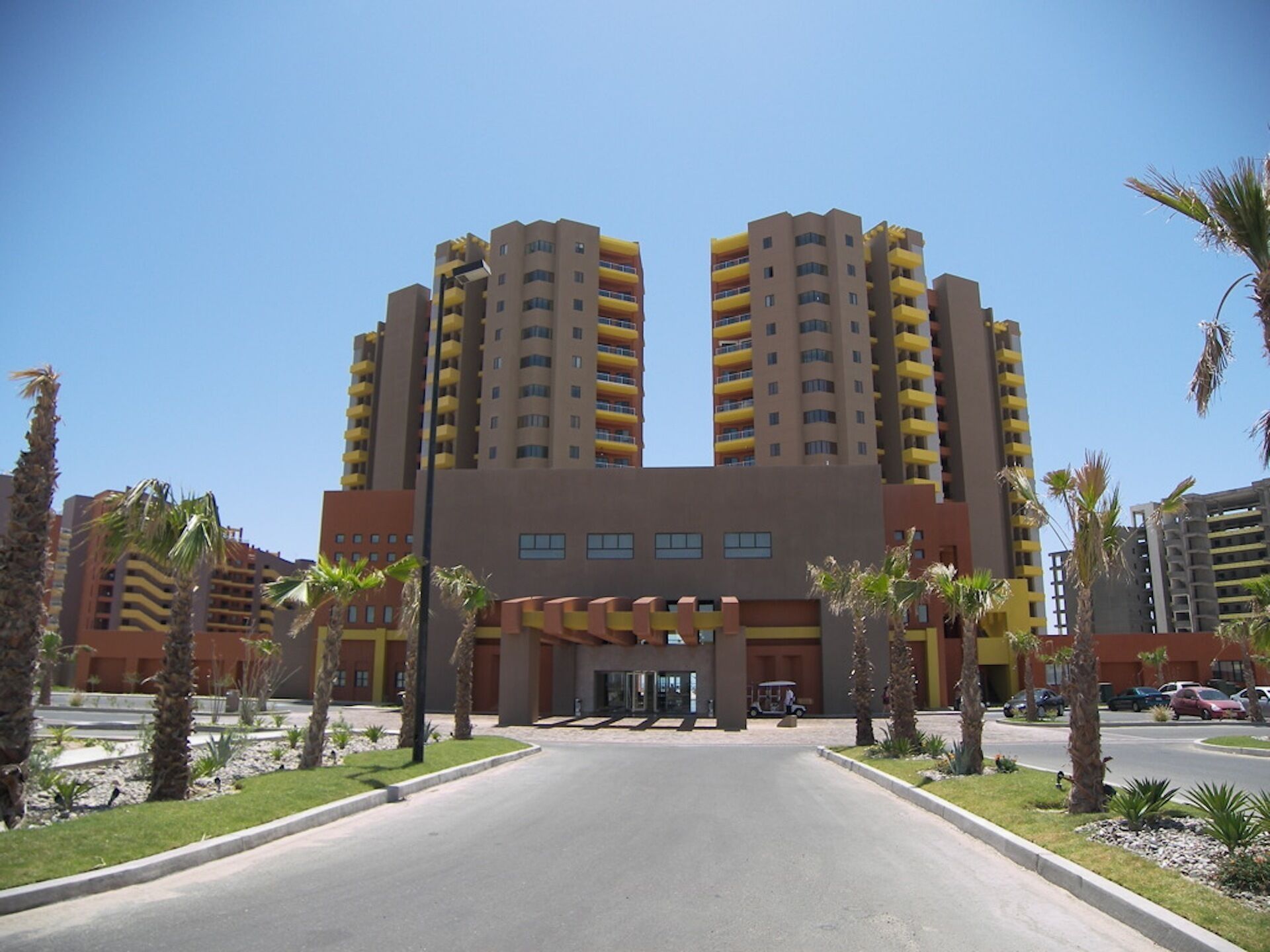 Een modern hotel met een centrale ingang, meerdere verdiepingen en een parkeerplaats.