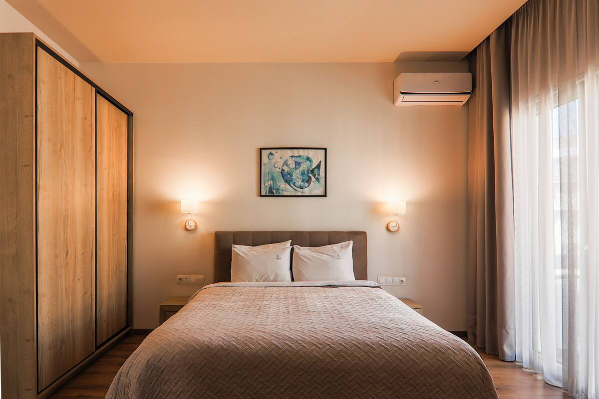 A bedroom with a large bed, wooden wardrobe, two bedside tables, a wall-mounted air conditioner, and a window with curtains.