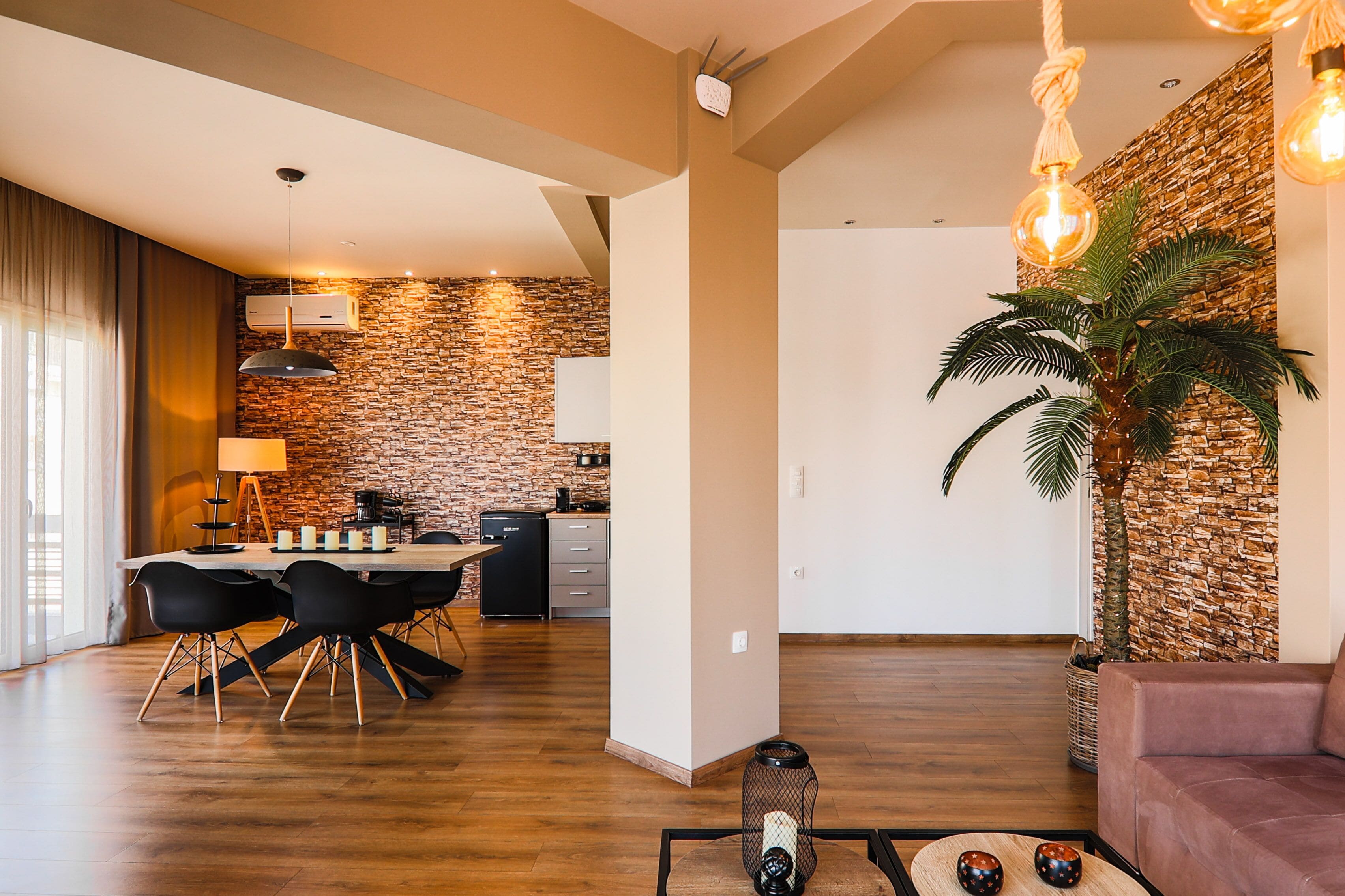 Una sala moderna con piso de madera, un comedor con mesa y sillas, una cocina con pared de piedra y una planta grande en maceta.