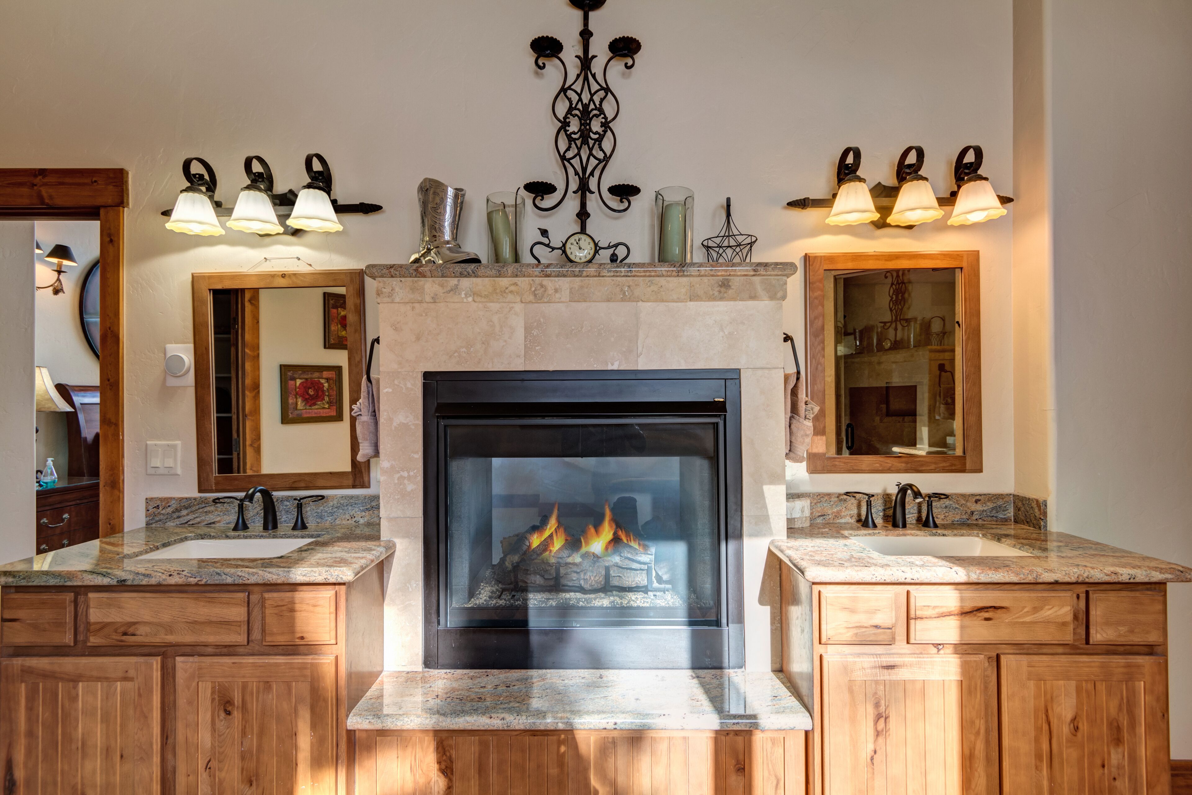 A bathroom with a fireplace, wooden cabinets, granite countertops, and wall-mounted lighting fixtures.