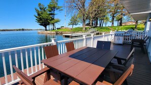 A deck with a table and chairs overlooking a calm lake, with boats docked nearby and a grassy area with trees.