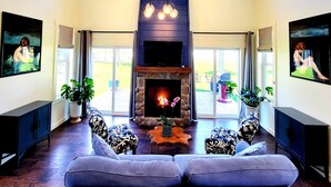 A spacious living room with a stone fireplace, a large sofa, and two armchairs. There are potted plants and a TV mounted on the wall.