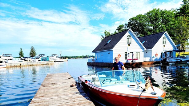 A wooden dock extending into calm water with a red boat moored at the end. In the background, there is a white house with a dark roof and a blue window.