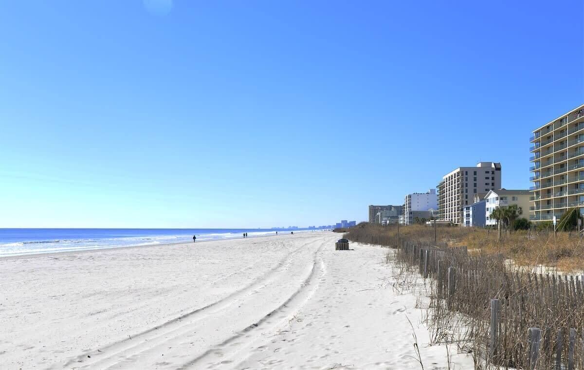 A beach with a sandy path leading to a building with multiple floors.