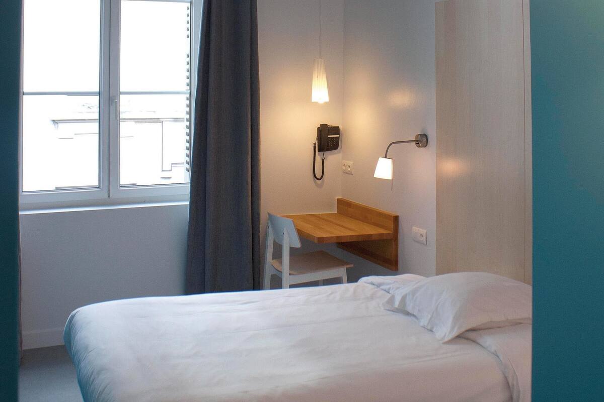 Una habitación de hotel con cama, escritorio, silla, teléfono y ventana con cortinas.