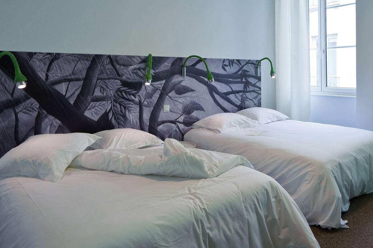 Habitación de hotel con dos camas, un mural grande de ramas de árboles y una ventana con luz natural.