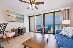 A living room with a wooden table, a blue sofa, a ceiling fan, a TV, and a balcony with a view of the ocean.
