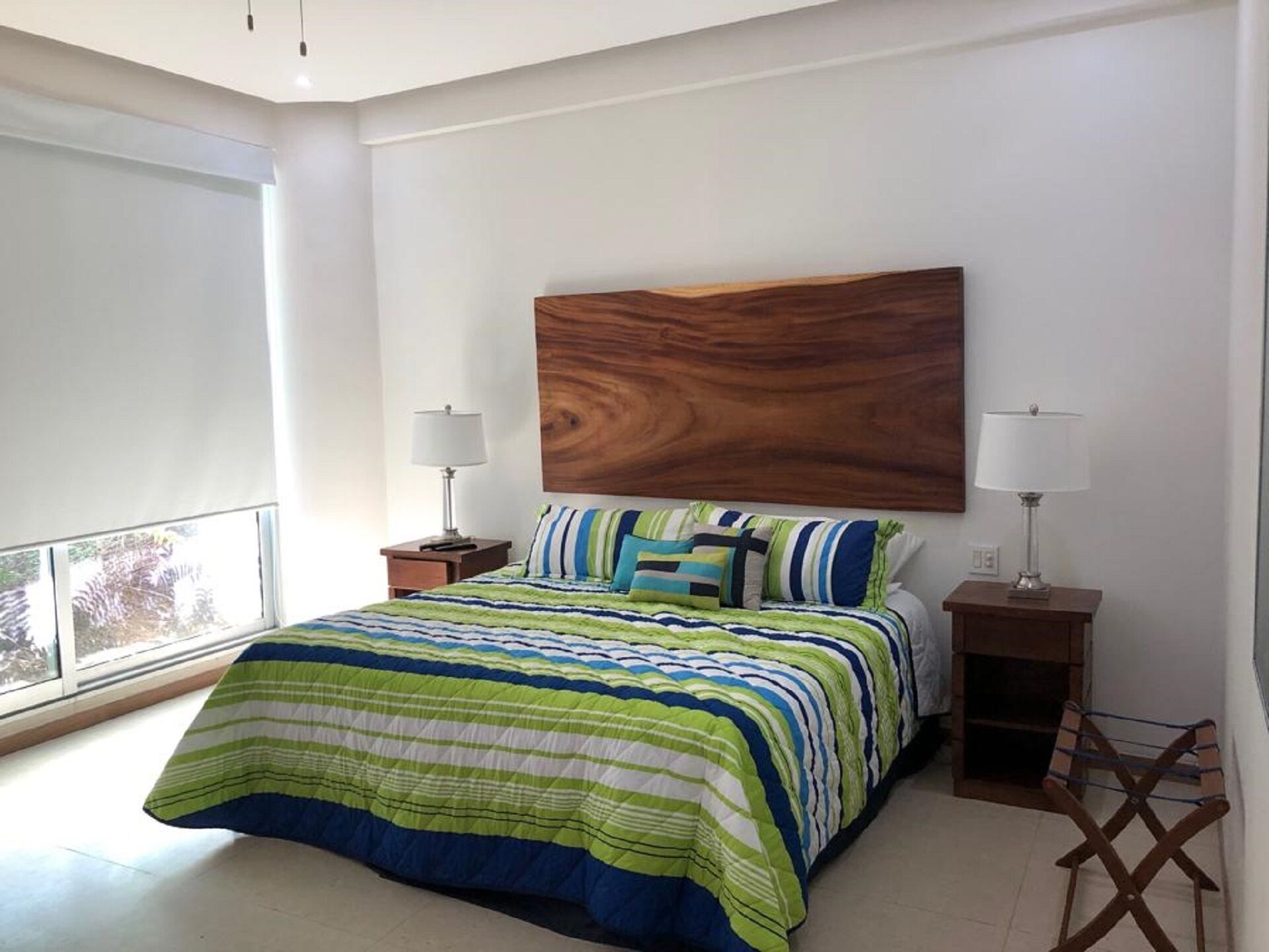 Une chambre à coucher avec un grand lit, deux tables de chevet avec des lampes, une tête de lit en bois et une vue sur l’extérieur par une fenêtre.