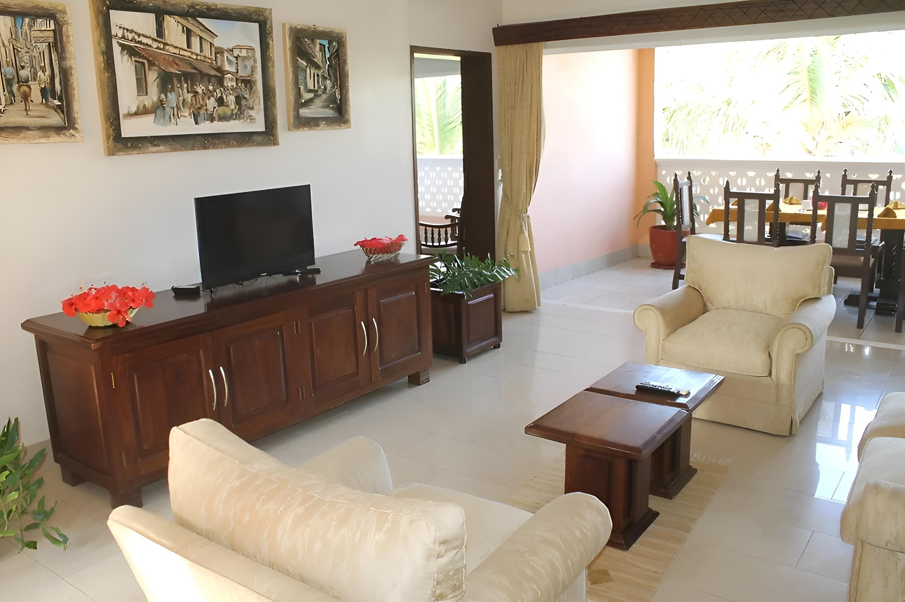 A spacious living room with a flat-screen TV, a wooden cabinet, and a beige sofa. There are two framed pictures on the wall and a dining area with a table and chairs visible through an open door.