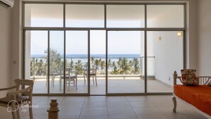 Une pièce spacieuse dotée d’une grande porte-fenêtre coulissante donnant sur un balcon avec vue sur l’océan et les palmiers.