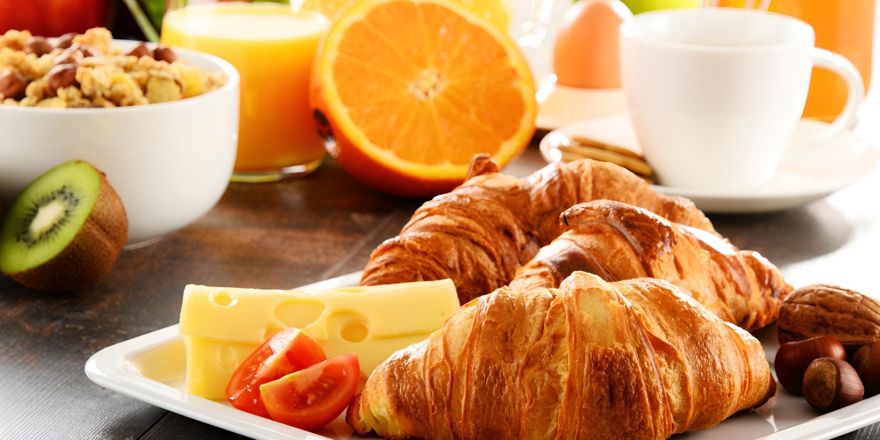A breakfast setting with croissants, cheese, tomatoes, kiwi, orange juice, and a cup of coffee.