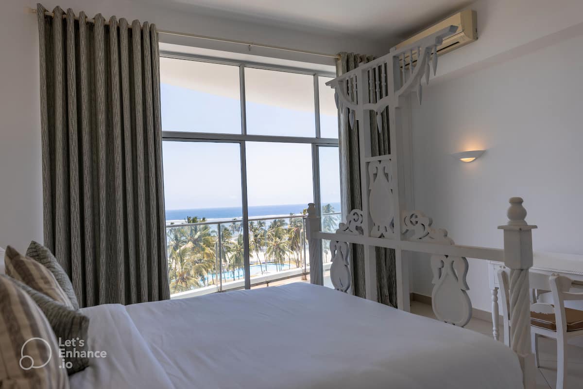 A bedroom with a large window, a bed, a white nightstand, and a view of the ocean.