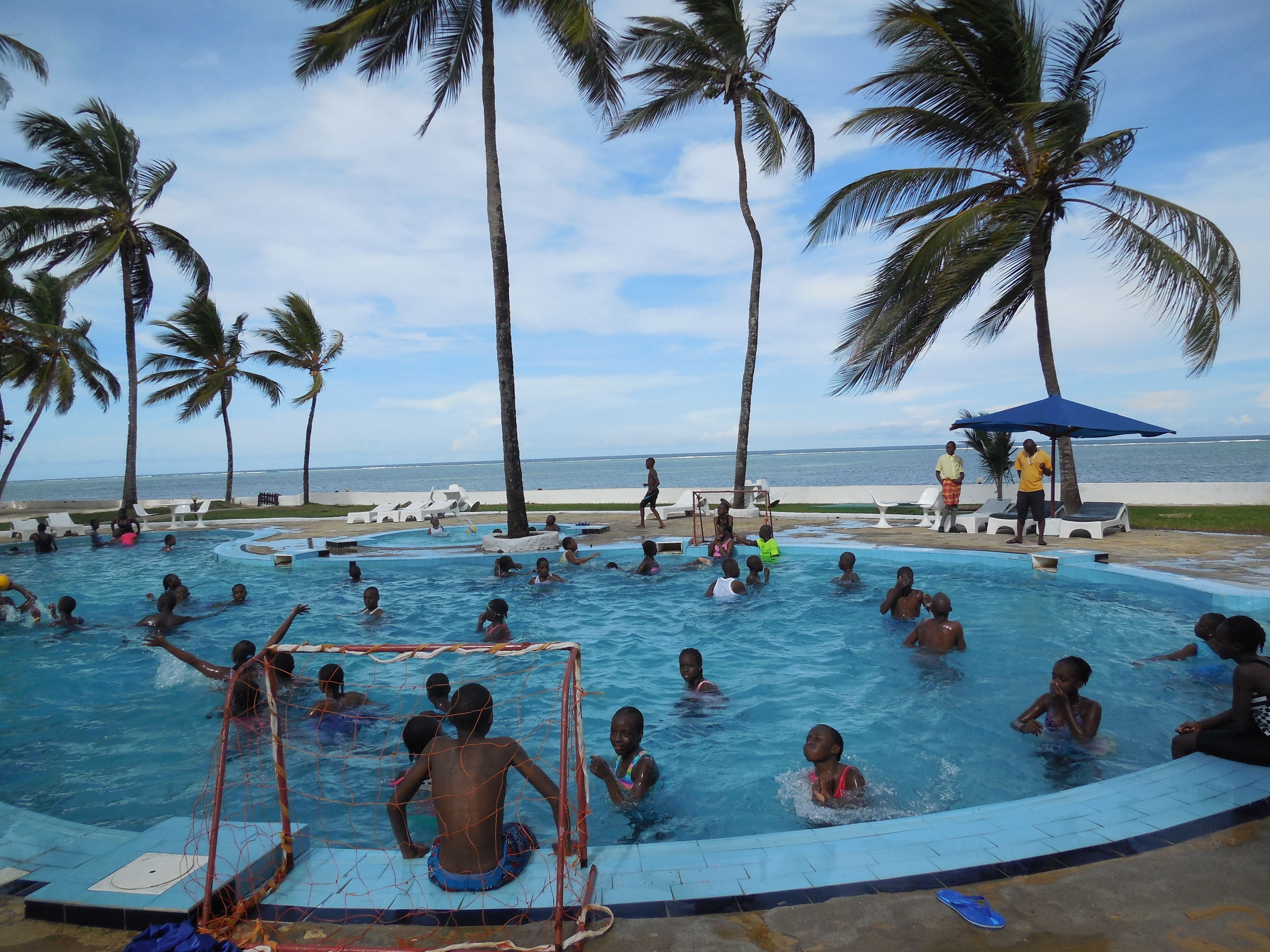 Zona de piscina con varias personas nadando y jugando, rodeada de palmeras y vistas al mar.