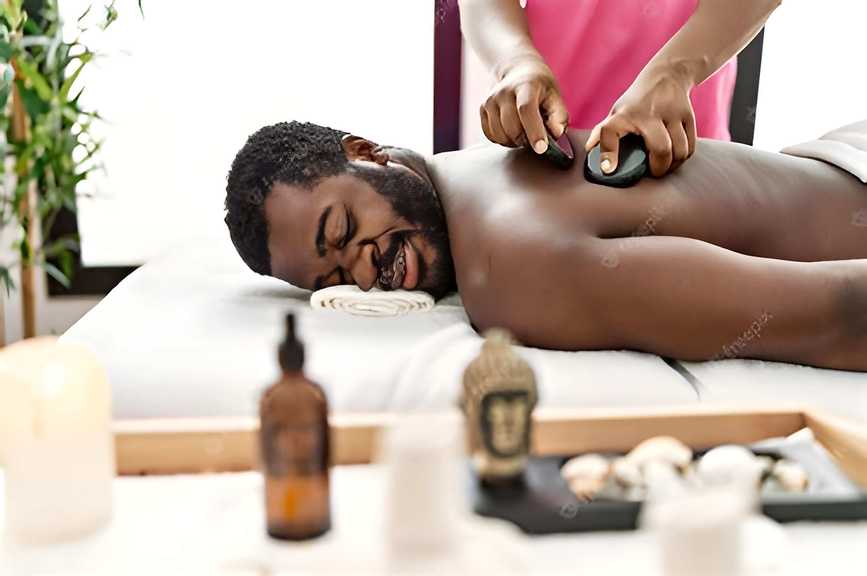 A person receiving a massage with a massage tool on a massage table, surrounded by candles, a small Buddha statue, and a bottle of essential oil.
