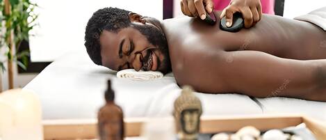 A person receiving a massage with a massage tool on a massage table, surrounded by candles, a small Buddha statue, and a bottle of essential oil.