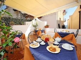 Un área de comedor al aire libre con mesas dispuestas para el desayuno, mostrando platos de pasteles y una jarra de jugo.