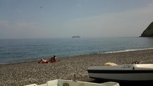 Una playa de piedrecitas con dos personas tomando el sol, un barco llamado 'SWIZZ' en primer plano y un crucero a lo lejos.