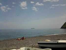 Una playa de piedrecitas con dos personas tomando el sol, un barco llamado 'SWIZZ' en primer plano y un crucero a lo lejos.