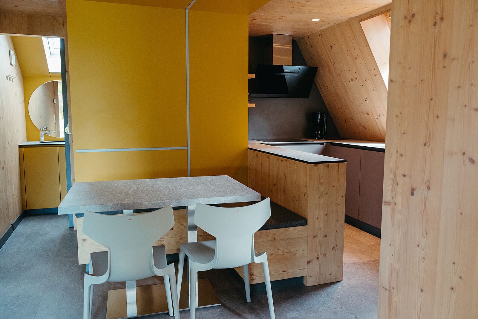 Een kleine keuken met een houten aanrecht, twee witte stoelen en een gele accentmuur.