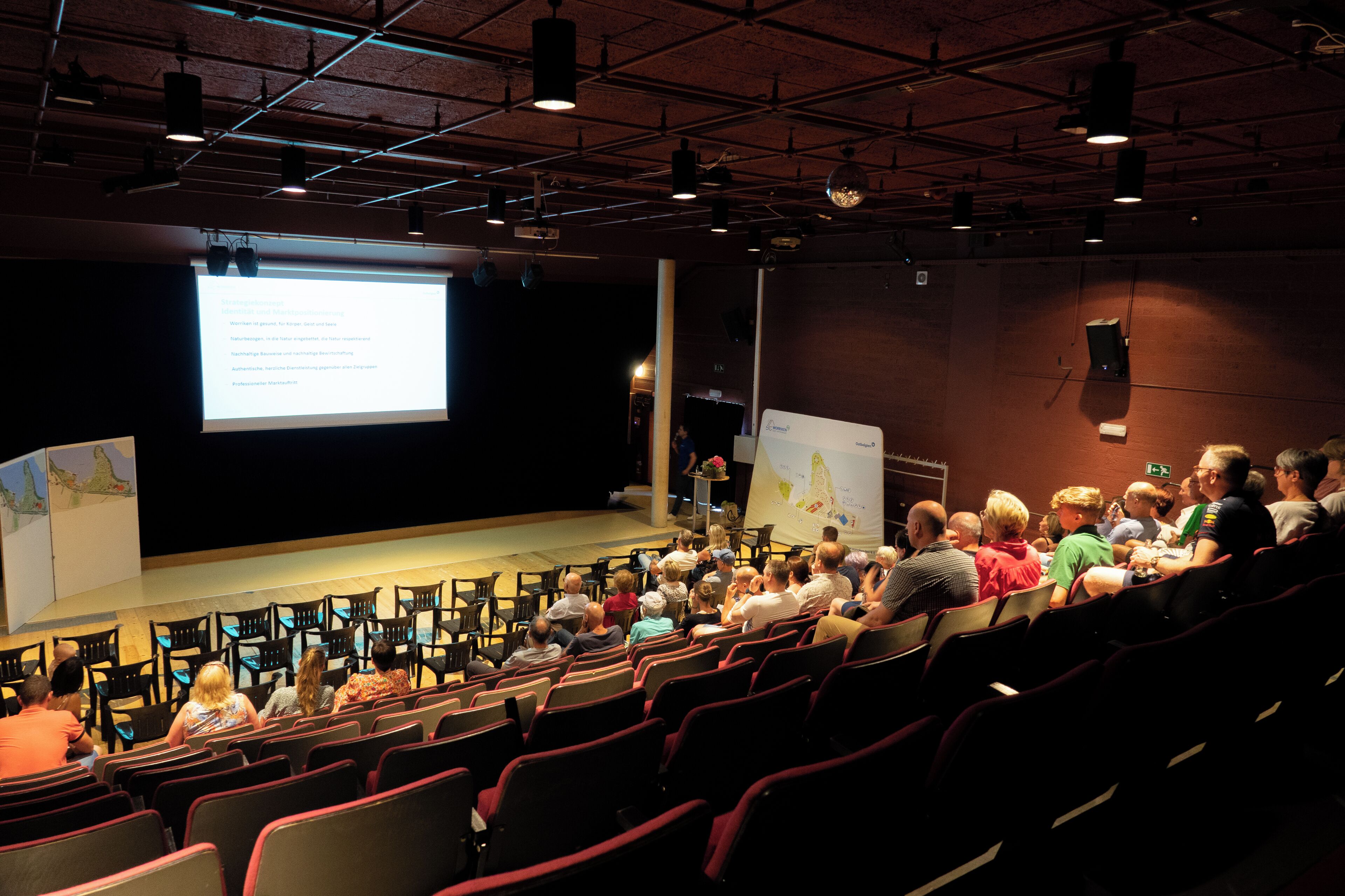 An auditorium with rows of seats, a stage, and a presentation screen.