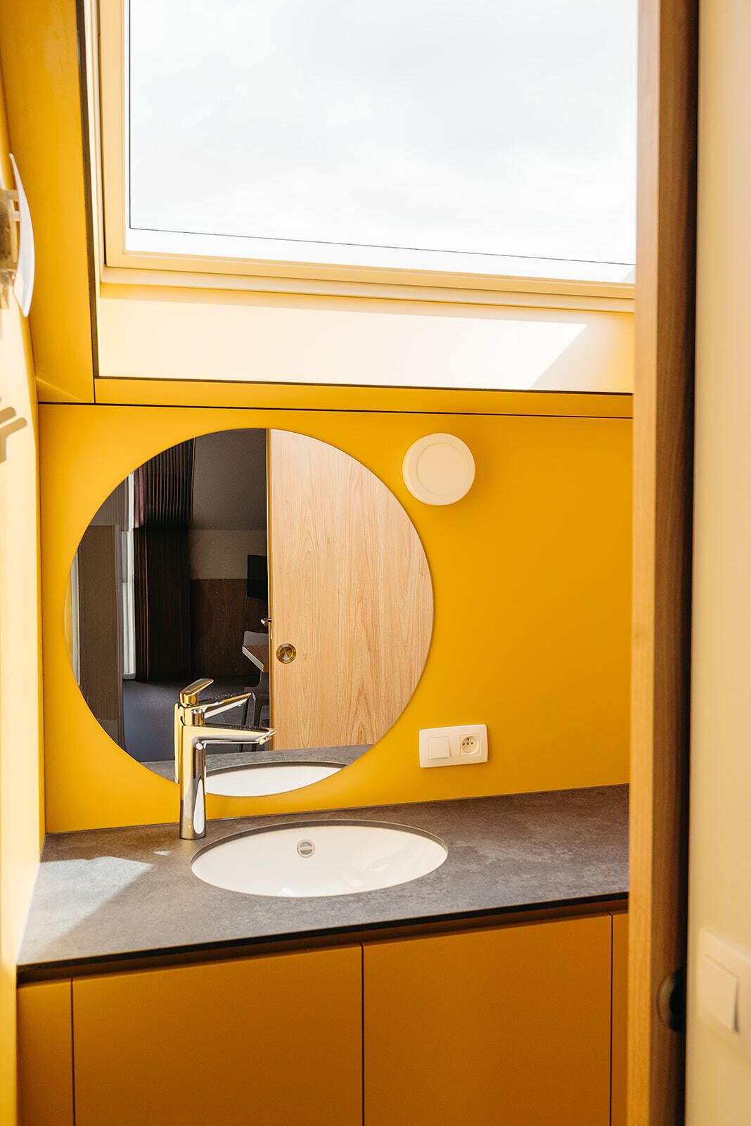 A bathroom with a yellow sink, a round mirror, and a window above the mirror.
