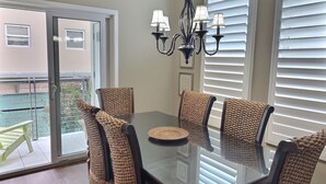 A dining area with a glass table surrounded by woven chairs, a chandelier, and white window blinds. - Fresh look at Shore Pleasure a family fav, steps from beach and downtown (Avila Beach)