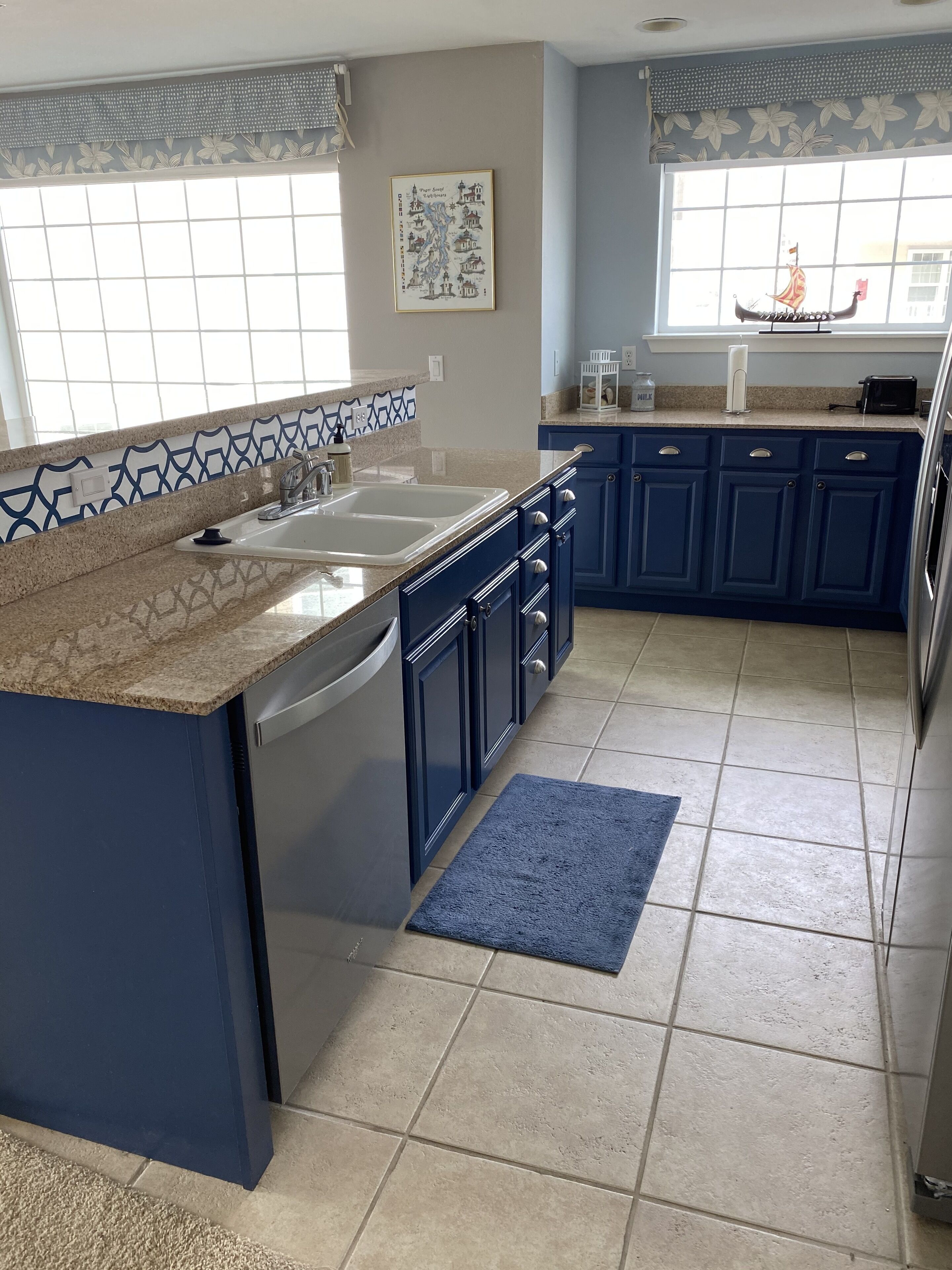A kitchen with blue cabinets, a granite countertop, a dishwasher, and a window with a decorative curtain.